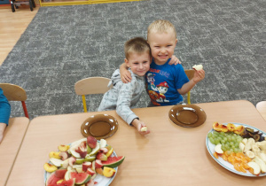 Dzieci siedzą wspólnie przy stole i degustują owoce: jabłka, banany, śliwki, brzoskwinie, mandarynki